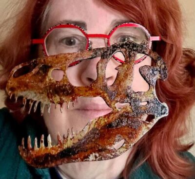 Picture of a red-haired woman with red glasses and a cut out of a dinosaur in front of her face.